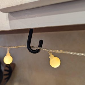 Könnte beinhalten: Ein schwarzer Metallhaken, der an einem weißen Schrank hängt, mit einer Lichterkette aus weißen Lichtern mit warmweißen Glühbirnen.