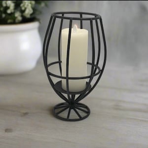 Puede incluir: Candelero de metal negro con diseño de jaula y una vela blanca de pilar en su interior. El candelero tiene una base redonda.
