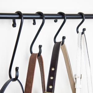 May include: Four black S-shaped hooks hang from a black metal rod. Three leather straps hang from the hooks.