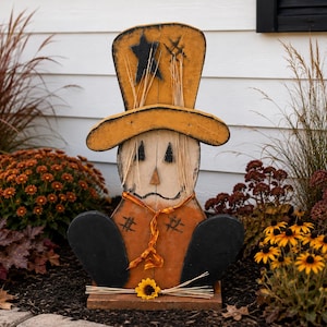 May include: A wooden scarecrow cutout featuring a tall yellow hat with a black star, a white face with triangular eyes, a small orange nose, and a stitched mouth. The body is orange with black patches, black arms, and a small sunflower accent.