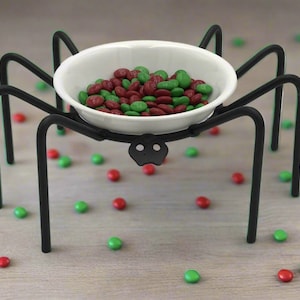 Op de afbeelding: Een zwarte metalen spinnenstandaard met een witte keramische kom gevuld met rode en groene snoepjes.