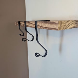 Può includere: Tre ganci in metallo nero montati su uno scaffale in legno. I ganci sono curvi e hanno un design decorativo.