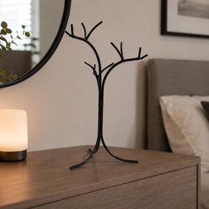 May include: A black metal jewelry tree with a tree branch design stands on a wooden dresser. The jewelry holder has multiple branches for hanging necklaces and bracelets. A white lamp and a vase with greenery are also on the dresser.