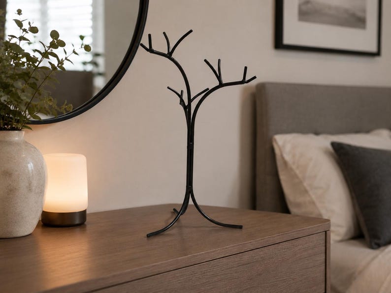 May include: A black metal jewelry tree with a tree branch design stands on a wooden dresser. The jewelry holder has multiple branches for hanging necklaces and bracelets. A white lamp and a vase with greenery are also on the dresser.