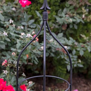 May include: Black metal garden trellis with a pointed top and a circular ring in the middle. The trellis is standing in a garden with pink and white flowers.