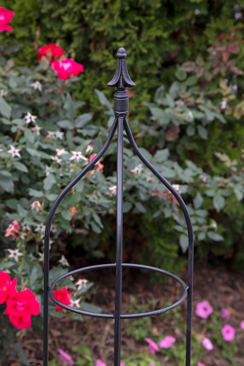 May include: Black metal garden trellis with a pointed top and a circular ring in the middle. The trellis is standing in a garden with pink and white flowers.
