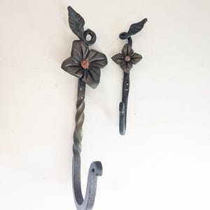 May include: Two handcrafted metal wall hooks. One hook features a twisted stem and a flower with a copper center. The other hook has a simpler design with a four-petal flower. Both have leaf accents and a dark, rustic finish.