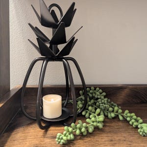 May include: A black metal candle holder with a unique, layered design. The holder features a small, lit candle inside and is accented by a green succulent plant. The candle holder is on a wooden surface.