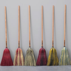 May include: Six upright brooms with wooden handles and colorful bristles. The bristles are in shades of red, cream, green, yellow, and blue. The brooms are arranged against a gray wall.