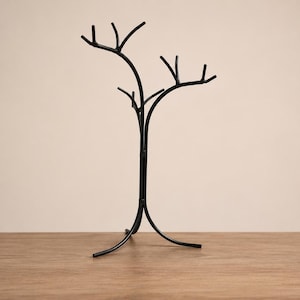 May include: A black metal tree-shaped decorative stand. The stand has a central trunk with multiple branches extending upwards. The base has three curved legs. The stand is on a wooden surface.