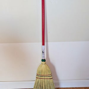 Könnte beinhalten: Ein traditioneller Besen mit rotem Holzstiel und natürlichem Strohkopf. Der Besenkopf ist mit grüner, roter und schwarzer Schnur befestigt. Der Besen steht aufrecht vor einer weißen und beigefarbenen Wand.