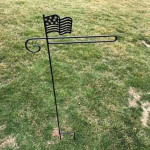 May include: A black metal flag holder with a decorative scroll design. The holder is designed to hold a small flag, and it is shown with a miniature American flag.