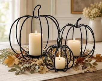 May include: Three black metal wire pumpkin-shaped candle holders of varying sizes, each containing a single white pillar candle. The wire frames are constructed from curved metal strips forming a cage-like structure with a stem at the top.