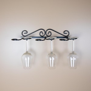 May include: Black metal wine glass rack with four glass stems hanging from it. The rack has a decorative scroll design.