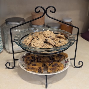May include: A black metal tiered stand with two glass plates. The top plate holds a variety of oatmeal raisin cookies. The bottom plate holds a variety of chocolate chip bars.