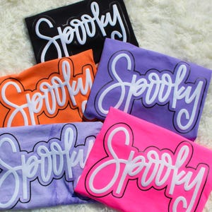 May include: A collection of folded t-shirts in various colors, including black, orange, purple, and pink. Each shirt features the word "Spooky" in a white, cursive font with a black outline, perfect for Halloween.