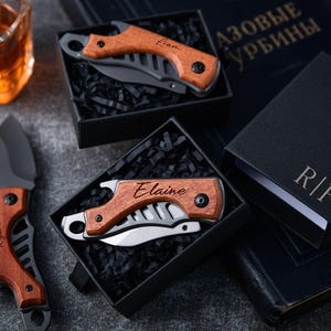 Puede incluir: Cuchillos de bolsillo con mangos de madera y nombres personalizados se exhiben en cajas de regalo negras. Un cuchillo está abierto, revelando una hoja plateada. Los nombres "Liam", "Elaine" y "Ryan" están grabados en los mangos. Un vaso de líquido ámbar está en el fondo.