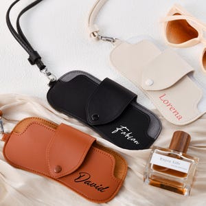 Puede incluir: Tres estuches de gafas de cuero personalizados con correas ajustables. Los estuches son marrón, negro y crema con los nombres "David", "Fabian" y "Lorena" escritos en ellos.