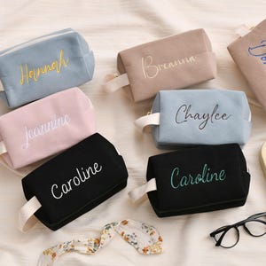 May include: A collection of personalized cosmetic bags in various colors, including blue, pink, black, and tan. Each bag features an embroidered name in a contrasting color, with a matching handle. A clock and glasses are also in the image.