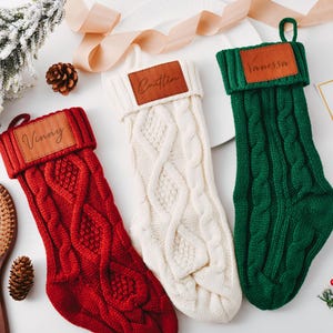 Puede incluir: Tres calcetines navideños de punto trenzado en rojo, crema y verde, cada uno con una etiqueta con el nombre de cuero sintético marrón. El calcetín rojo tiene "Vinny", el calcetín crema "Caitlin" y el calcetín verde "Vanessa".