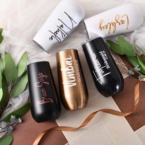 Puede incluir: Cinco copas de champán personalizadas con diferentes nombres y la palabra "dama de honor" en ellas. Las copas son de varios colores, incluyendo negro, blanco y dorado.