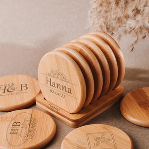 Puede incluir: Conjunto de seis posavasos redondos de bambú con diseños florales grabados y texto personalizado. Los posavasos están apilados en un soporte y presentan los nombres "Hanna" y "HB" con la fecha "02/04/22".