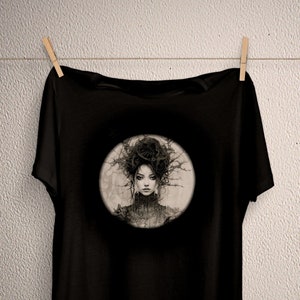 May include: Black t-shirt featuring a circular graphic of a woman with dark hair and a dark dress. The graphic is in shades of grey and white.