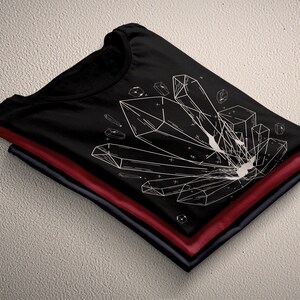 May include: A black t-shirt with a white geometric design of crystals. The shirt is folded and stacked on top of two other shirts, one red and one navy blue.