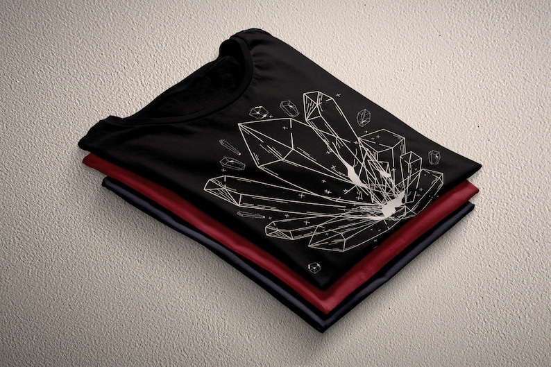 May include: A black t-shirt with a white geometric design of crystals. The shirt is folded and stacked on top of two other shirts, one red and one navy blue.