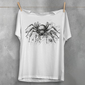 May include: White t-shirt featuring a detailed black and gray illustration of a large spider surrounded by smaller spiders. The graphic is centered on the shirt, creating a striking design. The shirt is hanging on a clothesline.