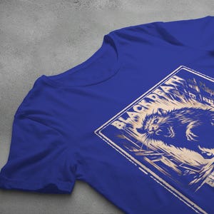 May include: A royal blue t-shirt with a vintage-style graphic of a snarling rat in front of a building. The graphic is in a light beige color and has the text "Black Death" in a stylized font.