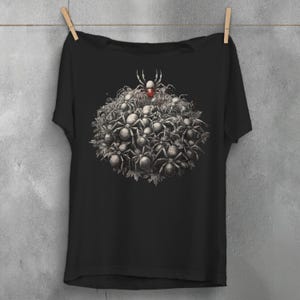 May include: Black t-shirt featuring a detailed illustration of a cluster of spiders. The design includes a central spider with a red marking, surrounded by a multitude of smaller spiders. The artwork is in grayscale, creating a striking contrast against the black fabric.
