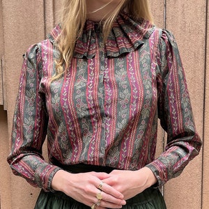 May include: A long-sleeved blouse with a ruffled collar and a striped pattern in shades of green, pink, and gold. The blouse has a floral print and is made of a soft, flowing fabric.