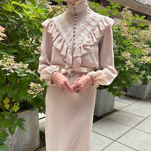 May include: A beige dress with a high ruffled collar and long sleeves. The dress has a lace trim and a satin ribbon tied at the waist.