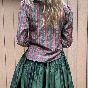 May include: A green ruffled skirt with a long-sleeved, patterned blouse. The blouse has a dark green, pink, and gold striped pattern with small floral details.