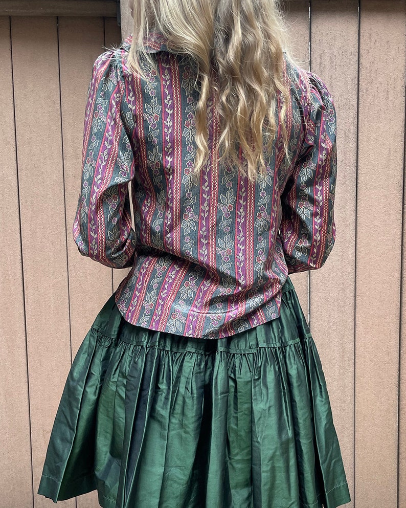May include: A green ruffled skirt with a long-sleeved, patterned blouse. The blouse has a dark green, pink, and gold striped pattern with small floral details.
