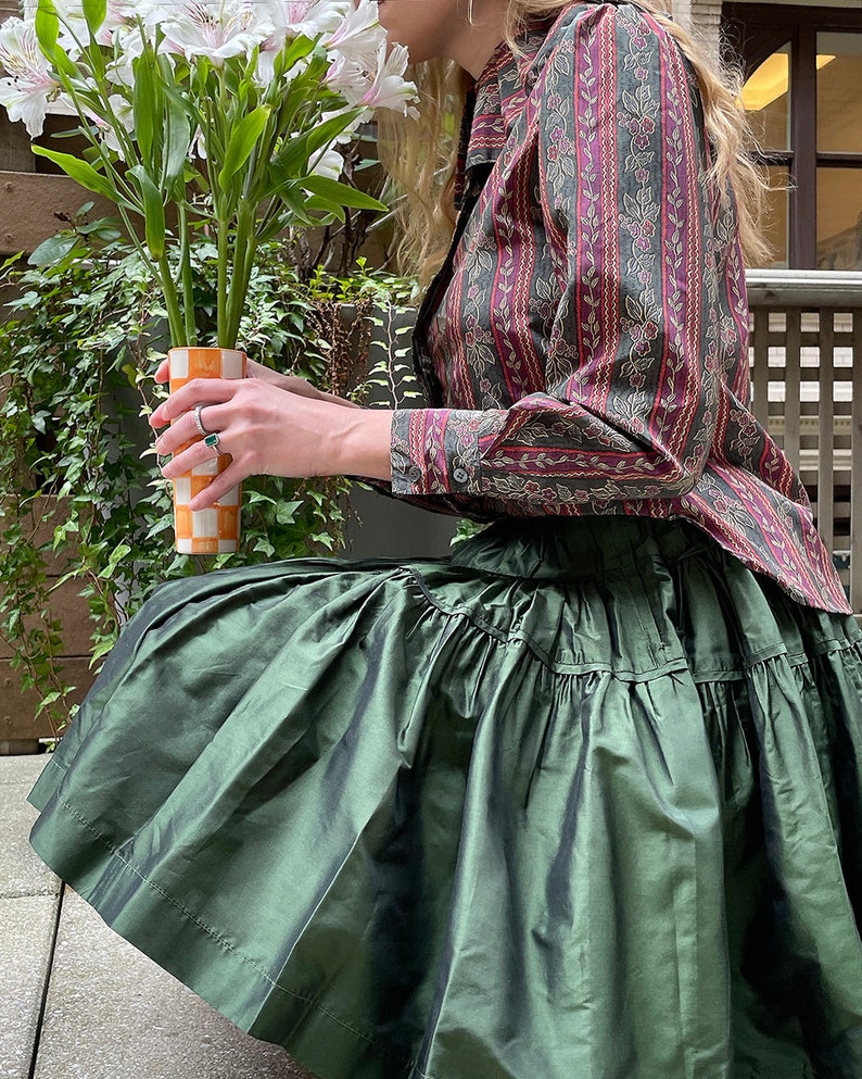 May include: A woman wearing a green, ruffled skirt and a long-sleeved, patterned shirt holds a vase with white flowers. The vase is orange and white with a checkered pattern.