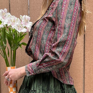 May include: A woman wearing a long-sleeved, patterned shirt with a green and pink floral design. The shirt has a ruffled collar and a button-up front. She is also wearing a dark green, tiered skirt.
