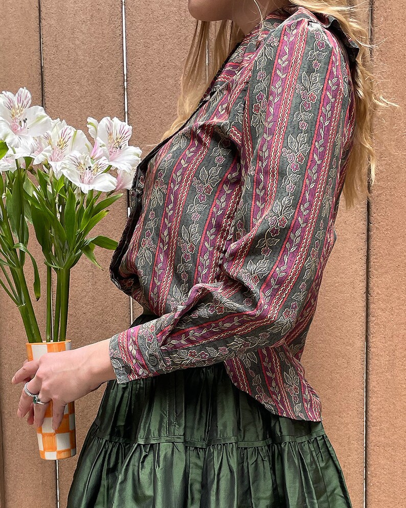 May include: A woman wearing a long-sleeved, patterned shirt with a green and pink floral design. The shirt has a ruffled collar and a button-up front. She is also wearing a dark green, tiered skirt.