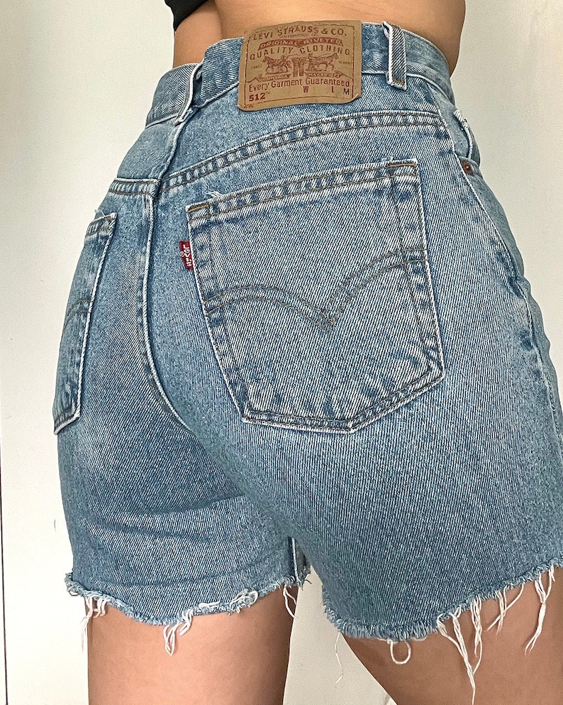 K&ouml;nnte beinhalten: Hellblaue Jeans-Shorts mit ausgefranstem Saum. Die Shorts haben eine Ges&auml;&szlig;tasche mit einem roten Levi's-Etikett und einem braunen Leder-Patch mit dem Levi's-Logo und dem Text "Original Riveted Quality Clothing" und "Every Garment Guaranteed 512 W L M".