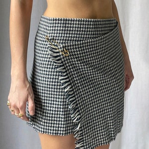 May include: A black and white houndstooth wrap skirt with a frayed hem and a gold safety pin detail.