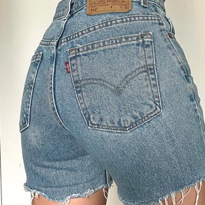 K&ouml;nnte beinhalten: Hellblaue Jeans-Shorts mit ausgefranstem Saum. Die Shorts haben eine Ges&auml;&szlig;tasche mit einem Levi's-Logo und ein braunes Leder-Etikett mit dem Levi's Strauss & Co.-Logo und dem Text "Original  Quality Clothing  Every Garment Guaranteed 512 W L M".