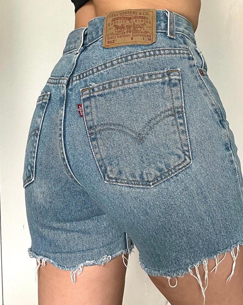 K&ouml;nnte beinhalten: Hellblaue Jeans-Shorts mit ausgefranstem Saum. Die Shorts haben eine Ges&auml;&szlig;tasche mit einem Levi's-Logo und ein braunes Leder-Etikett mit dem Levi's Strauss & Co.-Logo und dem Text "Original  Quality Clothing  Every Garment Guaranteed 512 W L M".