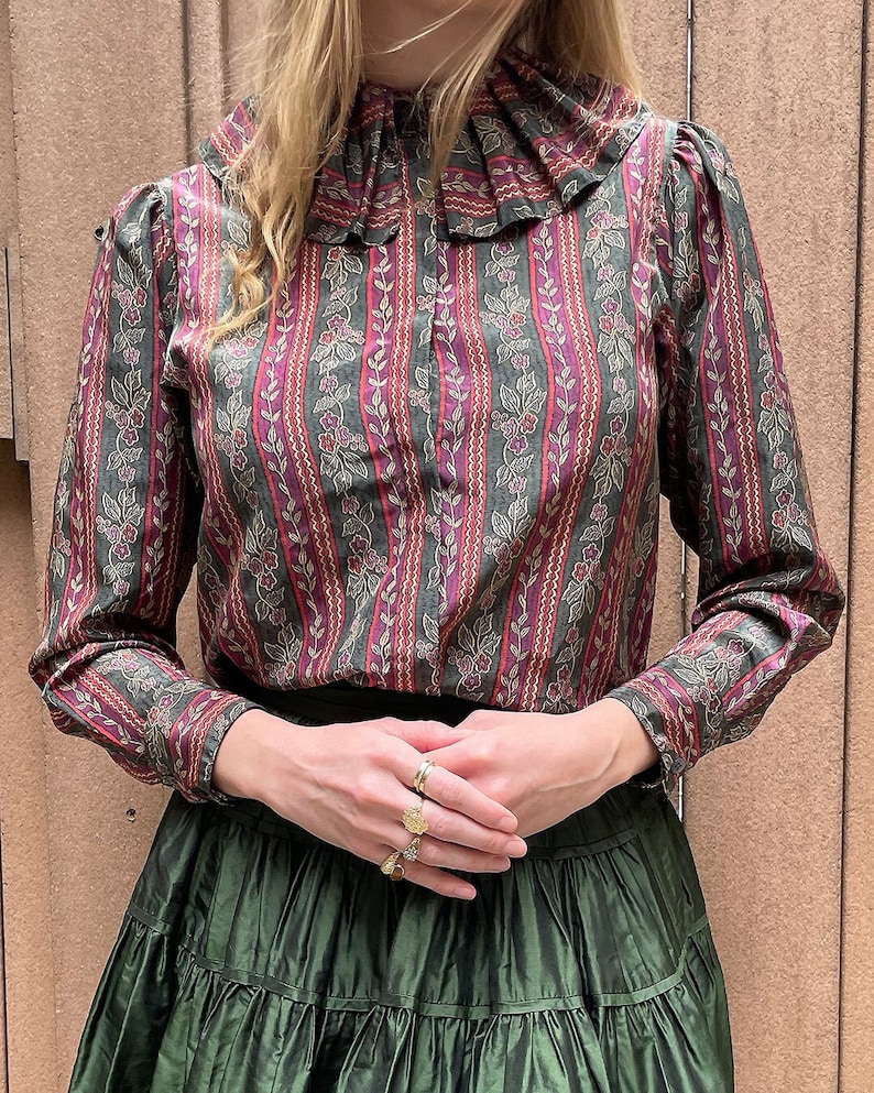 May include: A long-sleeved blouse with a ruffled collar and a striped pattern in shades of green, pink, and gold. The blouse has a floral print and is made of a soft, flowing fabric.