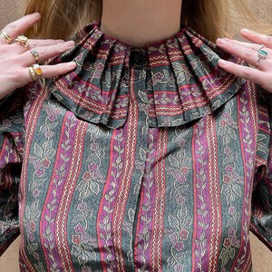 May include: A vintage-style blouse with a ruffled collar and a floral print. The blouse has a dark green background with pink and white floral accents.