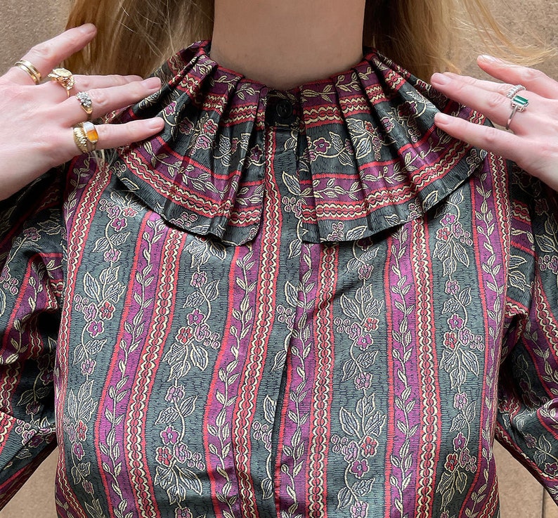 May include: A vintage-style blouse with a ruffled collar and a floral print. The blouse has a dark green background with pink and white floral accents.