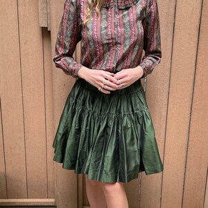 May include: A green tiered skirt with a floral print blouse. The blouse has a high collar with a ruffle.