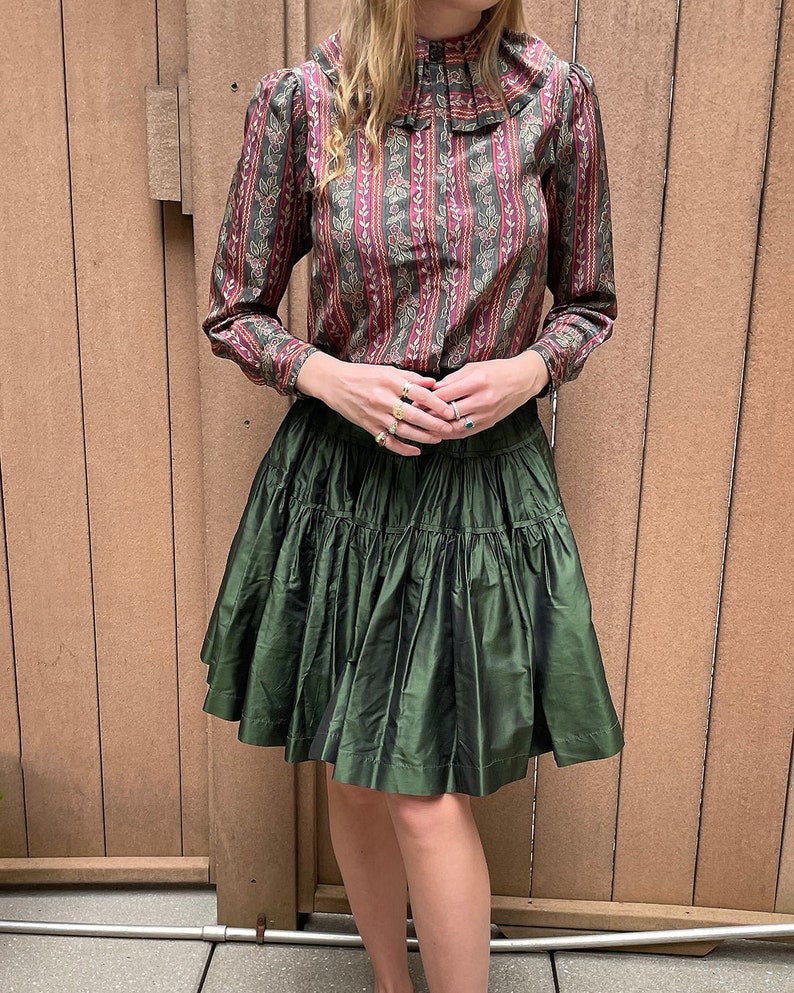 May include: A green tiered skirt with a floral print blouse. The blouse has a high collar with a ruffle.