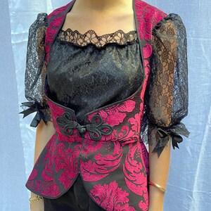 May include: A black and pink patterned vest with a black lace overlay. The vest has a cinched waist and a high neckline.