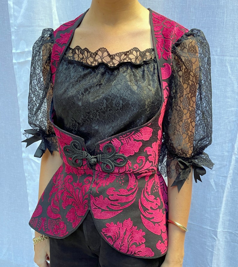 May include: A black and pink patterned vest with a black lace overlay. The vest has a cinched waist and a high neckline.